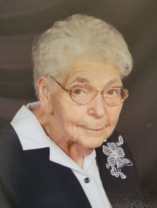 Portrait of Esther Borte, smiling warmly, wearing glasses and a navy cardigan with white floral embroidery.