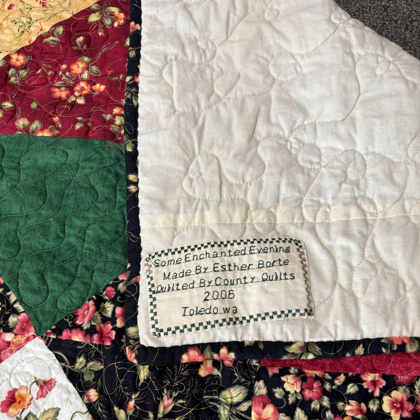 Corner of the showcase quilt, Some Enchanted Evening, with its hand-stitched label reading Made By Esther Borte, Quilted By County Quilts, 2006, Toledo WA — surrounded by burgundy, hunter-green, gold, and floral patchwork.